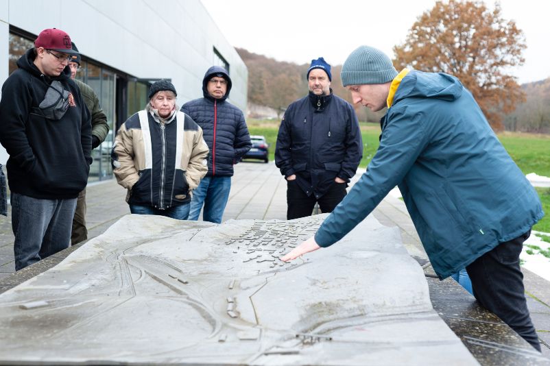 A memorial staff member begins a guided tour at the camp model. He points to one area of the model while visitors stand all around it.