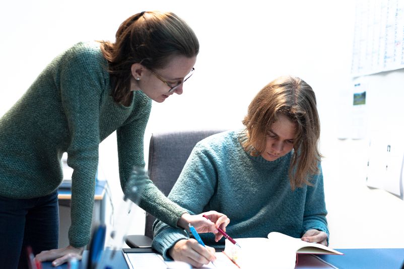 Two people at a table. The young man is sitting at a desk and looking at a book. The woman stands bent over the desk and points with a ballpoint pen at something in the said book.