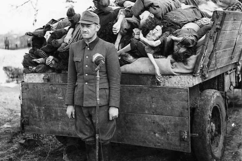 A man in uniform can be seen in front of a microphone. Directly behind him is a wooden trailer filled to the top with jumbled corpses. 