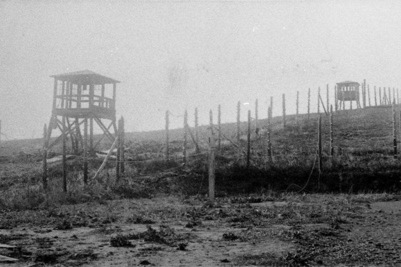 A wire fence runs up a slope. Next to it are two small, wooden watchtowers. 