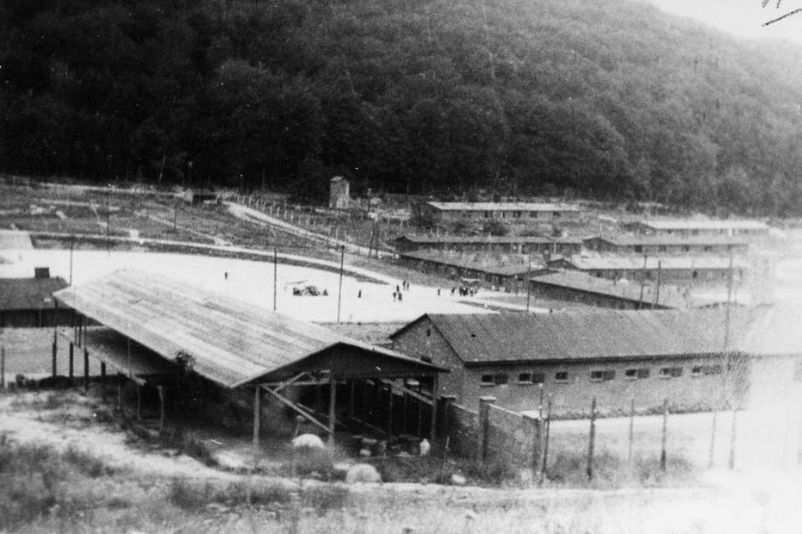 Blick aus südöstlicher Richtung auf den Appellplatz und den Lagereingang. Rechts im Hintergrund Baracken der SS. Im Vordergrund eine zum Holzhof gehörende Lagerhalle und daneben der Arrestzellenbau.