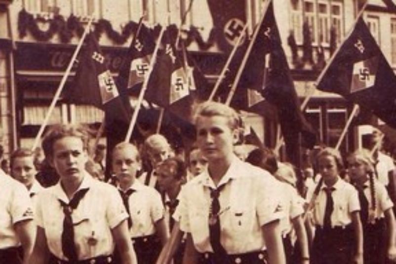 A photo of young women in uniform marching down a street in single file. They carry swastika flags. The houses on the roadside are also decorated with swastikas. 